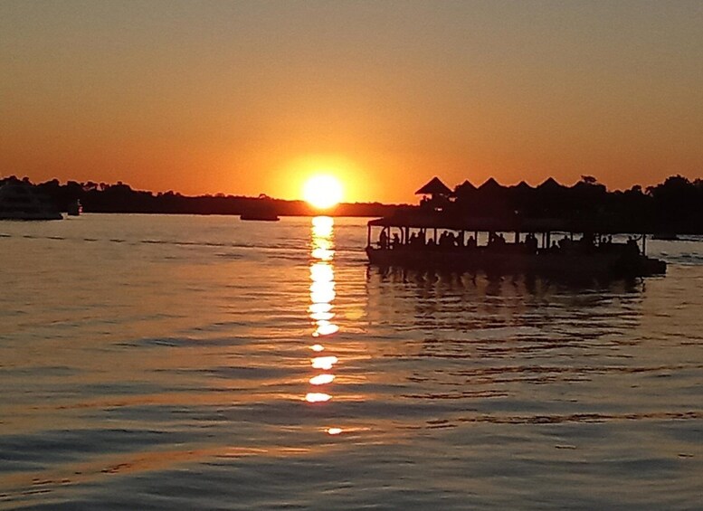 A sunset over a calm body of water with a boat and thatched-roof structures on the shore. 1