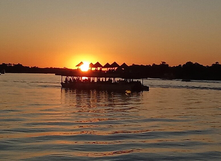 A sunset over a body of water with a boat carrying people and a structure with thatched roofs. 2