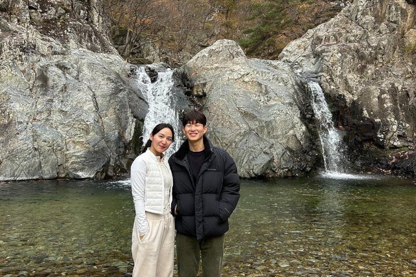 Two people standing in front of a waterfall with rocky terrain and a clear water pool. 5