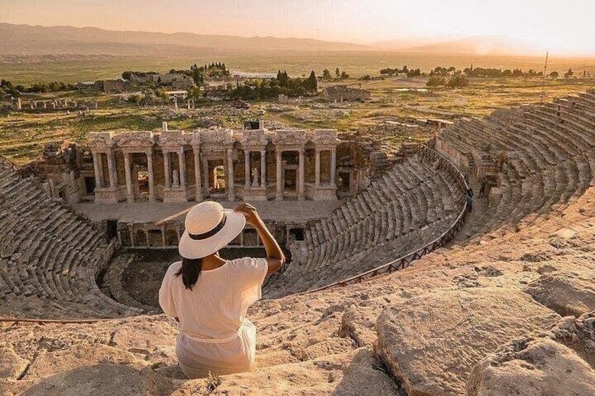 A person sitting on a sandy area overlooking an ancient amphitheater with ruins and a distant landscape. 2