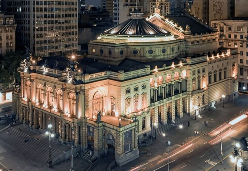 A grand, illuminated building with a dome and classical architecture, surrounded by a cityscape at night. 1
