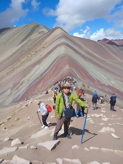 A hiker with a green jacket and a hat, trekking on a rocky path with a colorful mountain in the background. 5