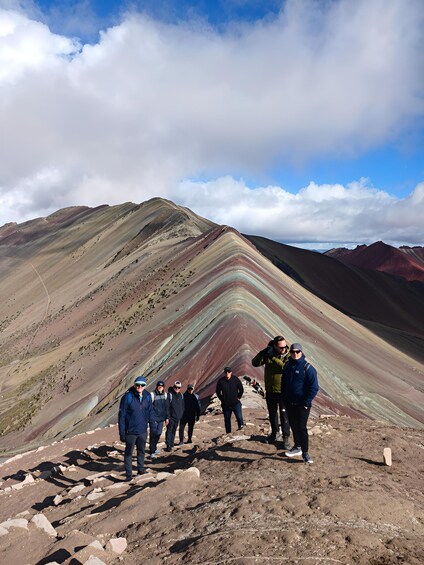 A group of people standing on a rocky mountain ridge with colorful stratified rock formations in the background. 2