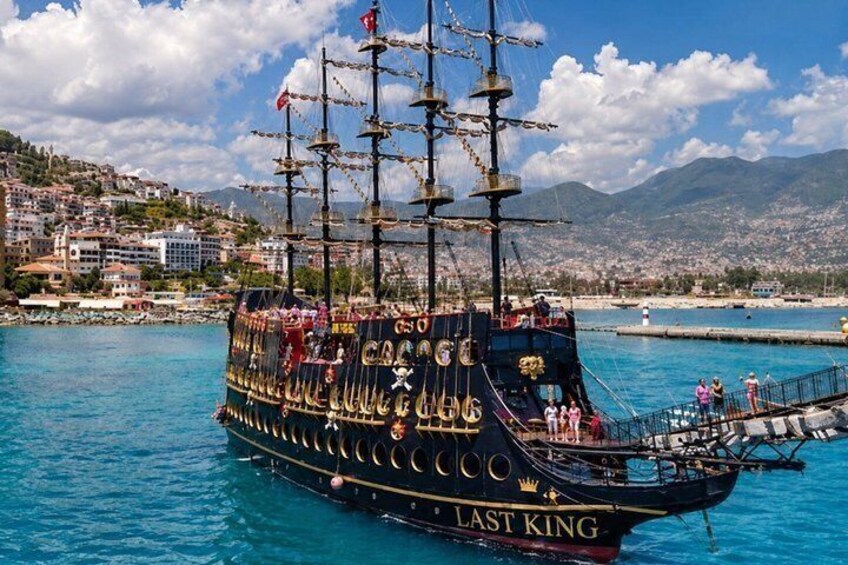 A tall ship named 'Last King' with multiple masts and flags, docked at a harbor with buildings and mountains in the background. 1
