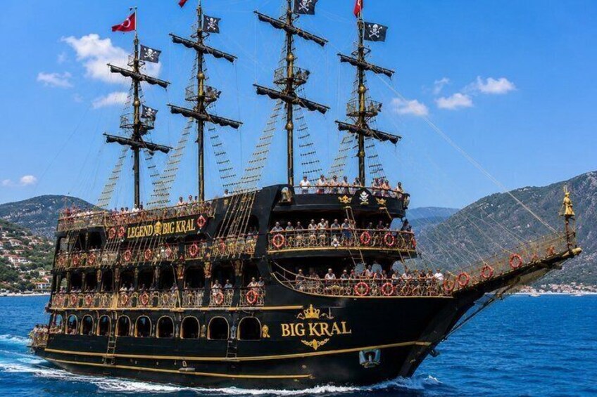 A large, ornate ship named 'Big Kral' with multiple decks and flags, sailing on a body of water with a mountainous coastline in the background. 3