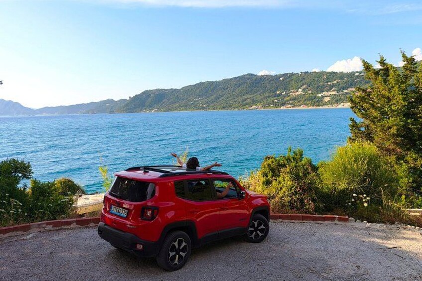 A red SUV parked on a gravel road with a scenic view of a blue lake and green hills in the background. 2