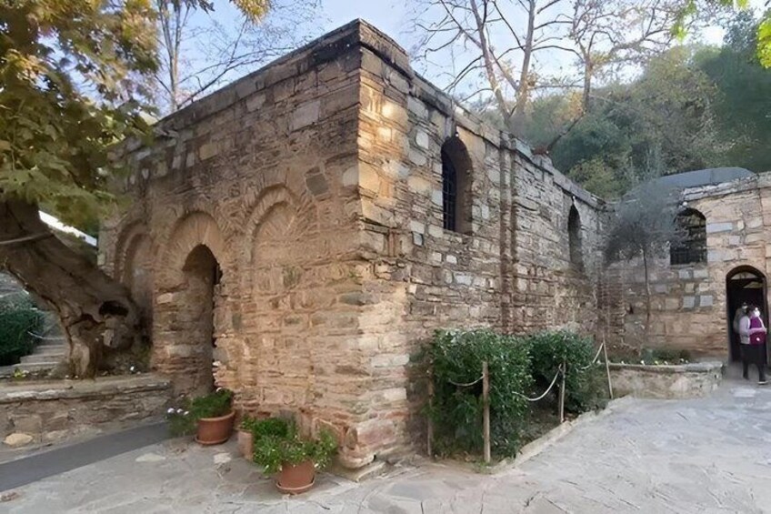 An ancient stone building with arched windows and a small door, surrounded by potted plants and a paved path. 3