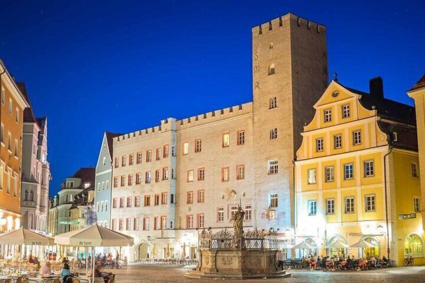 A historic building with a tower, a fountain, and surrounding cafes. 1