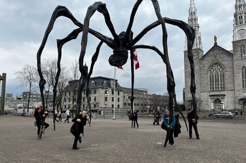 A large spider sculpture in an open plaza with people walking around, a church with Gothic architecture in the background. 5