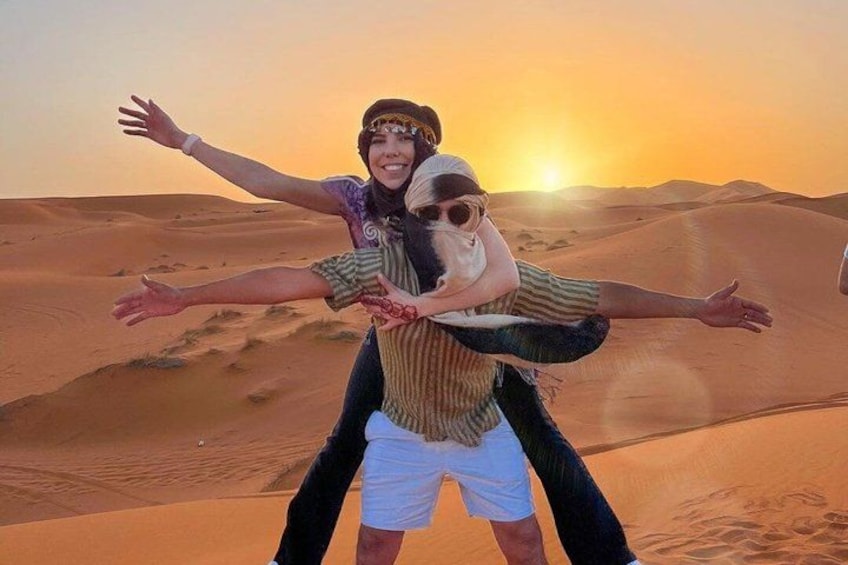 Dos personas se abrazan en un paisaje desértico con dunas de arena y un atardecer. 1