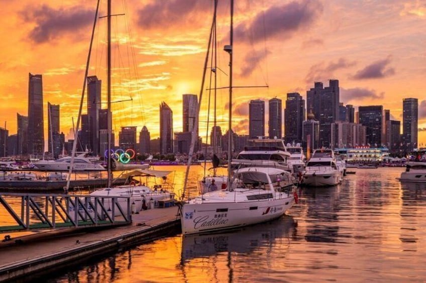 A marina with yachts, a city skyline, and the Olympic rings logo. 1