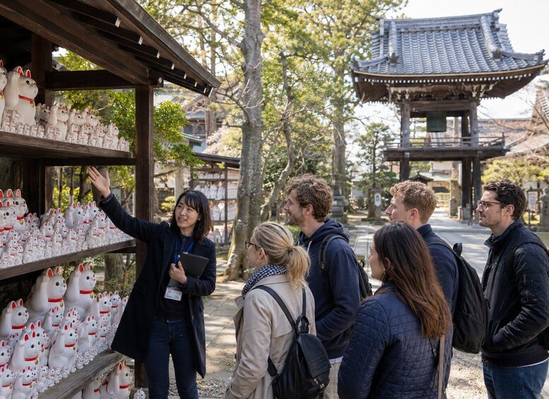 A group of people at a shrine with traditional Japanese architecture, including a pagoda and a shrine with a wooden structure displaying numerous Maneki-neko figurines. 1