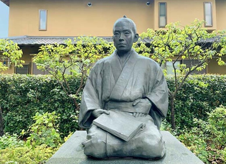 A statue of a seated figure in traditional attire, holding a book, with a building and greenery in the background. 2