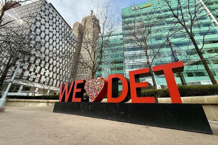 A sign with the words 'WE LOVE DET' and a heart shape in the middle, set against a backdrop of modern buildings. 4