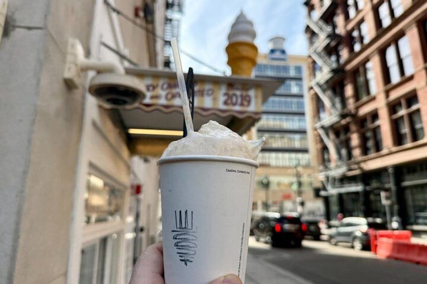 A hand holding a cup of iced coffee with a straw, a building with a sign that reads '2019', and a street with cars and buildings in the background. 2