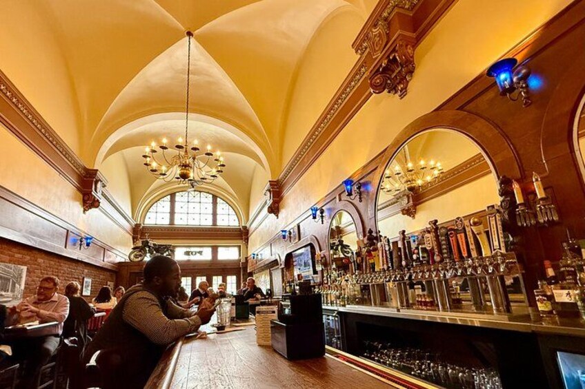 A bar with a high ceiling and arched windows, a bartender, and patrons seated at tables. 3