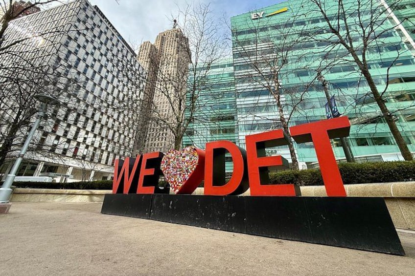 A sign with the words 'WE LOVE DET' and a heart shape in the middle, set against a backdrop of modern buildings. 3
