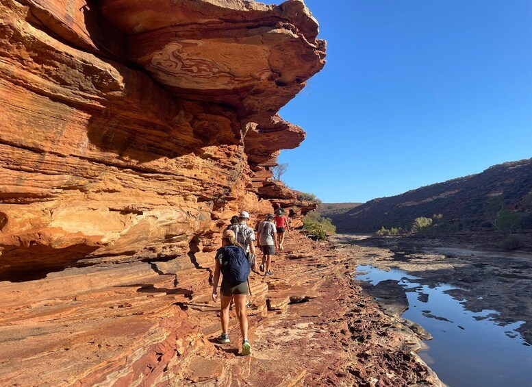 Hikers on a rocky riverbank with layered red rock formations. 5