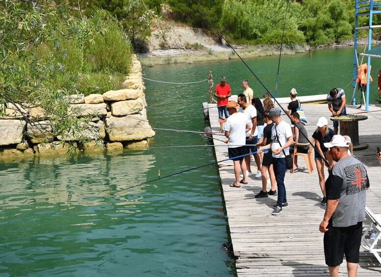 A group of people fishing on a wooden pier by a calm river with greenery in the background. 2