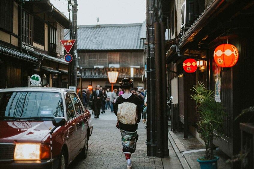A person walking down a narrow street lined with traditional buildings, a red car parked on the side, and red lanterns hanging above. 4