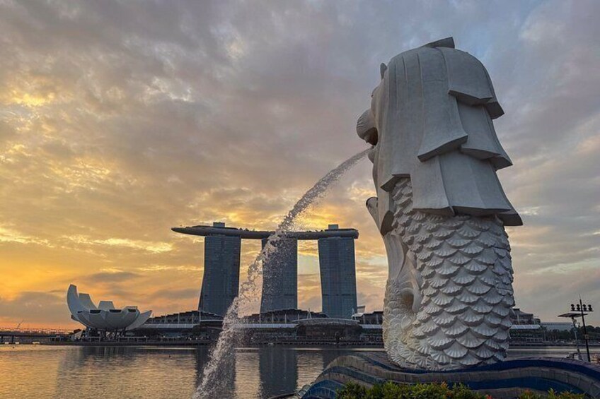 A large stone lion statue with water flowing from its mouth, a modern architectural building with a distinctive sail-shaped roof, and a sunset sky. 4