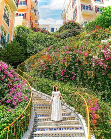 A woman in a white dress descending a colorful staircase surrounded by lush greenery and vibrant flowers. 1
