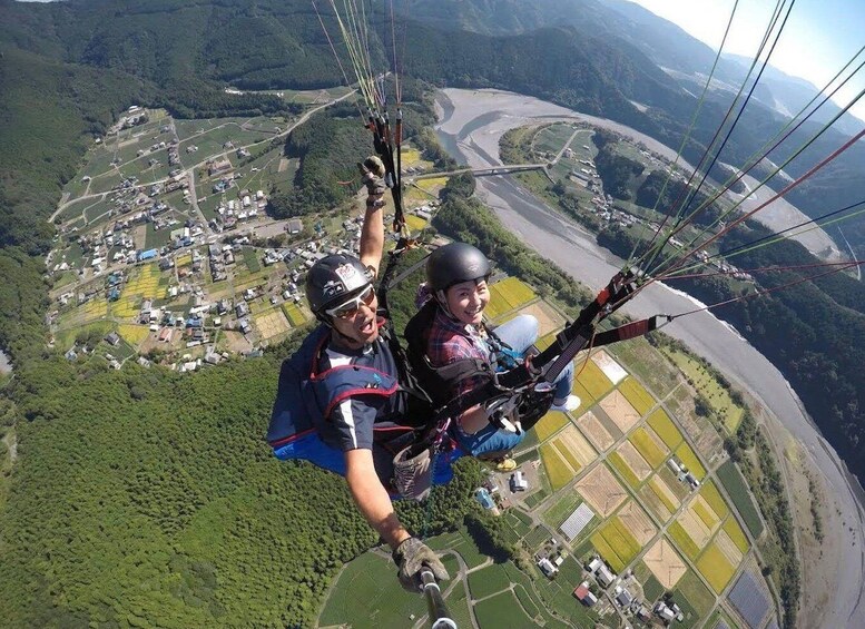 Two people paragliding over a landscape with fields, a river, and a bridge. 5