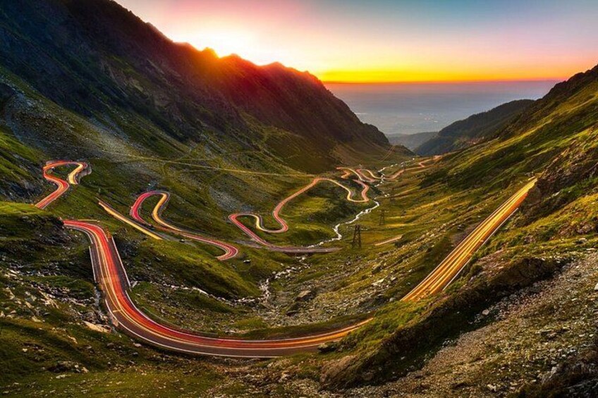 A winding mountain road with multiple curves, surrounded by lush green hills and a sunset in the background. 3
