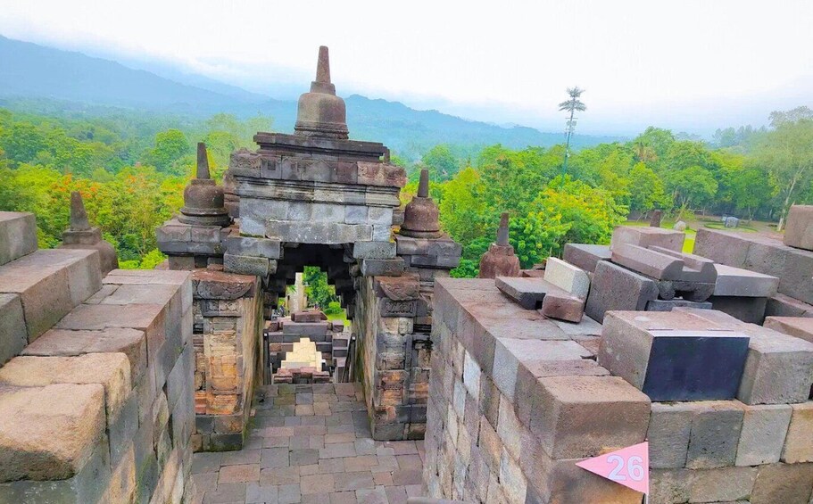 An ancient stone structure with a central passageway, surrounded by stone blocks and set against a lush green forest backdrop. 5