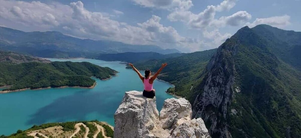 A person standing on a rock with arms outstretched, overlooking a turquoise lake and mountainous landscape. 1