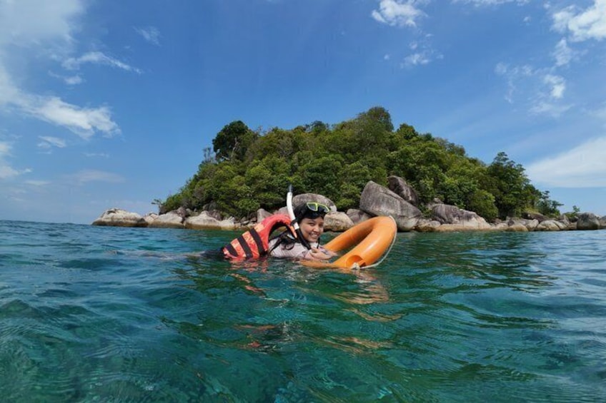 A person floating on water with a ring buoy, surrounded by clear blue sea and a rocky island with greenery. 5