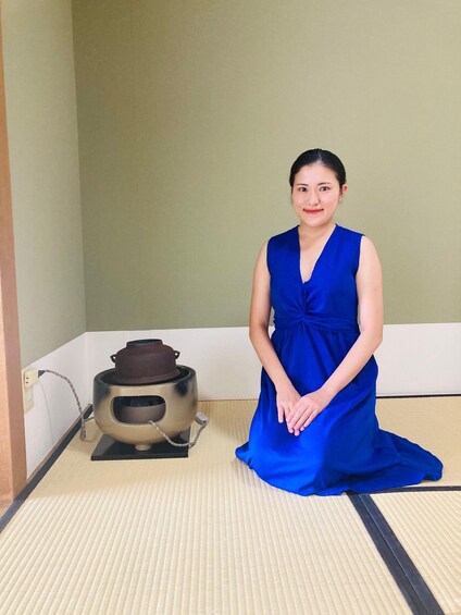 A person in a blue dress kneeling on tatami mat flooring next to a traditional Japanese tea kettle. 2