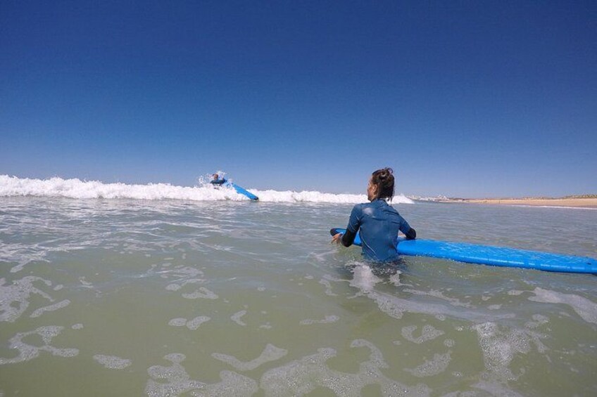 A person in a wetsuit holding a blue surfboard in the ocean, with waves and a clear sky in the background. 2
