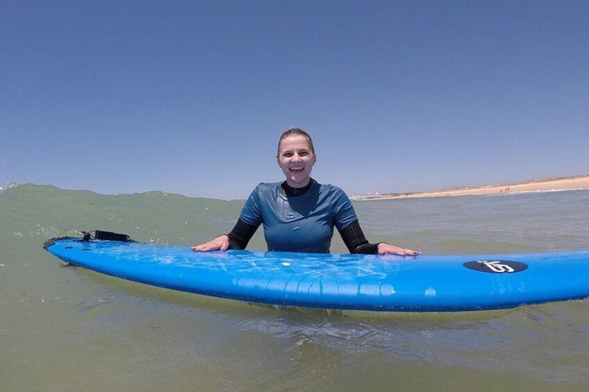 A person holding a blue surfboard with a logo, standing in shallow water with a beach and ocean in the background. 3