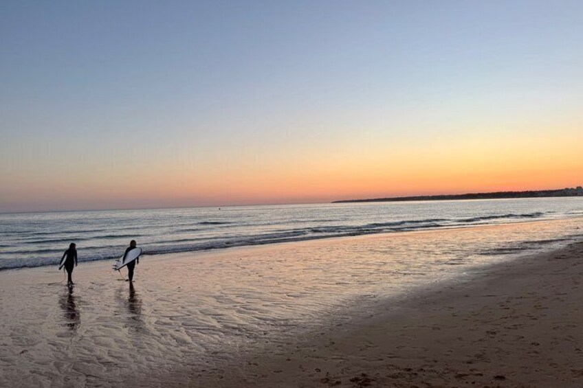 A beach at sunset with two people walking along the shore, one holding a surfboard. 2