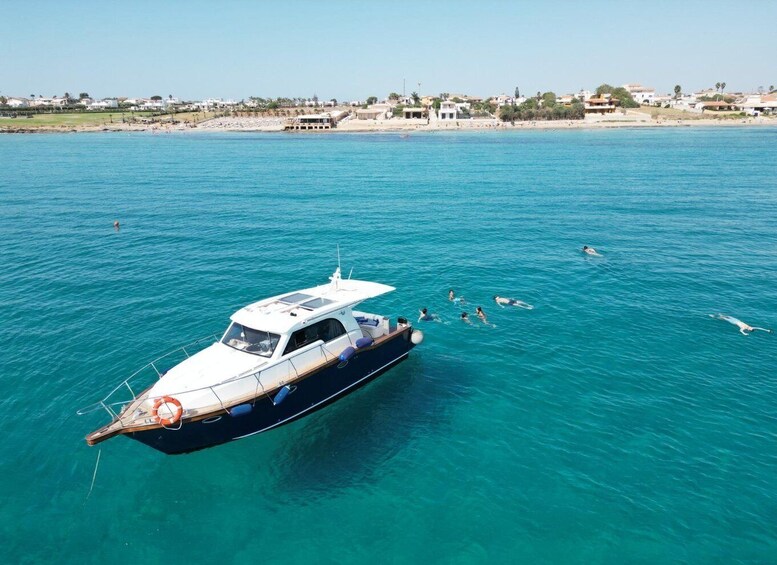 A boat on clear turquoise water with swimmers in the distance and a beach with buildings in the background. 1