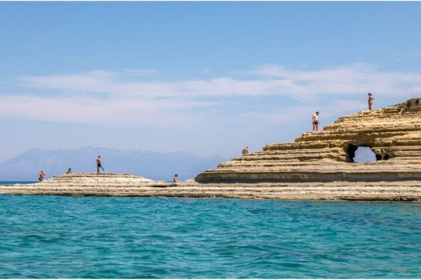 A rocky coastal landscape with people standing on the cliffs and a clear blue sea. 4