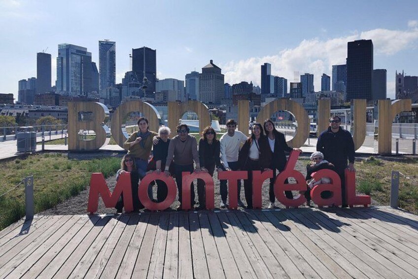 A group of people standing in front of a large sign with the words 'Bonjour Montréal' on a wooden boardwalk with a city skyline in the background. 4