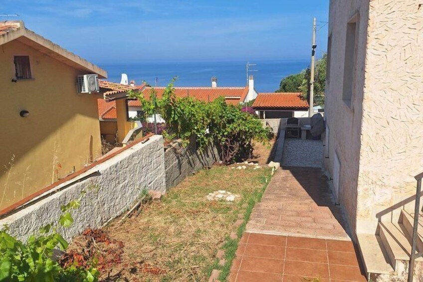 A narrow stone pathway leading to a building with a tiled roof, surrounded by greenery and houses with red-tiled roofs. 5