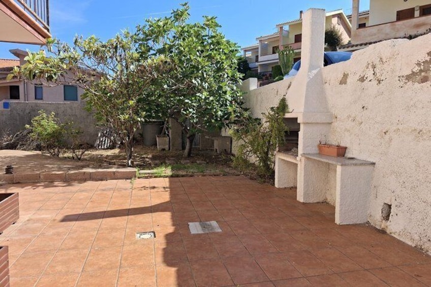 A courtyard with tiled flooring, a tree, and a white wall with a chimney. 4