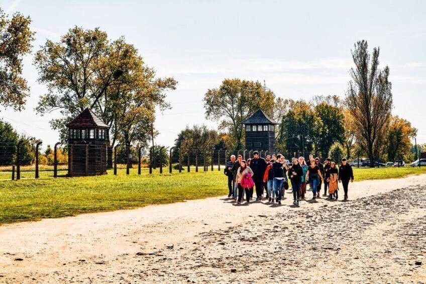 A group of people standing on a dirt path with a fence and two wooden structures in a park. 1