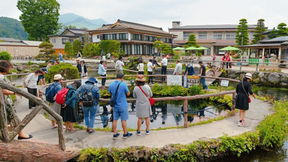 A group of people standing by a pond with a wooden bridge, surrounded by traditional buildings and greenery. 4