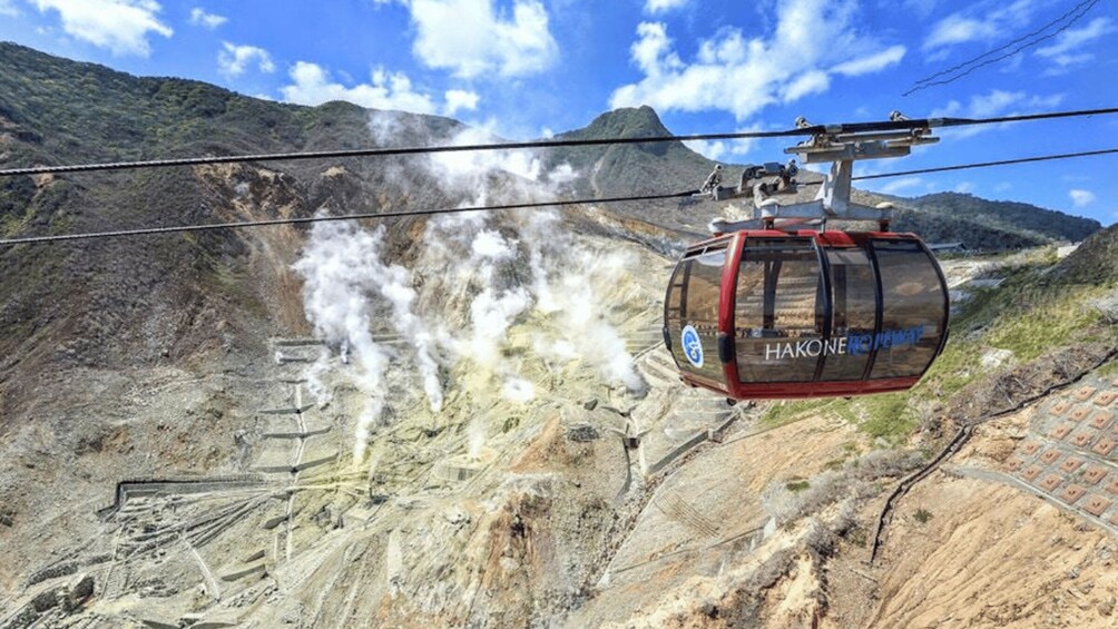 A cable car with 'HAKONE ROYAL' branding, suspended over a mountainous landscape with visible steam or smoke, and a clear blue sky. 2