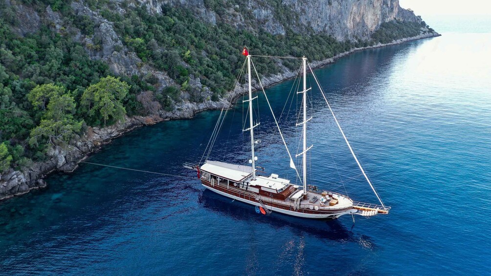 A sailboat anchored in a clear blue sea with a rocky coastline and dense greenery in the background. 5