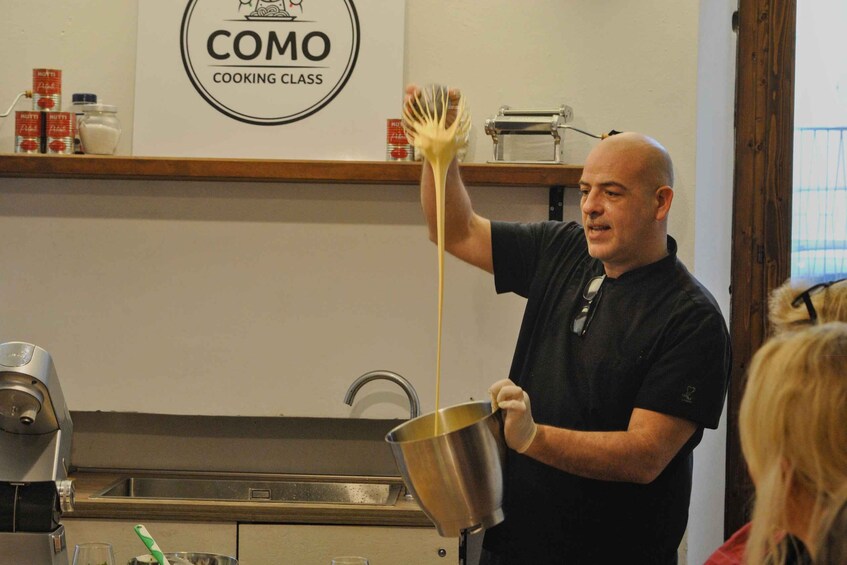 A person holding a pot of liquid, possibly cooking, in a kitchen with a 'COMO Cooking Class' sign. 5