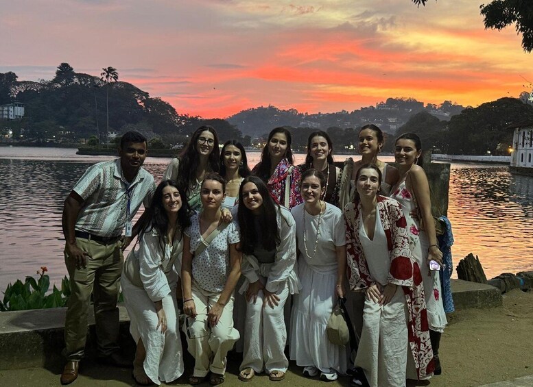 A group of people posing by a lake at sunset with a cityscape in the background. 1