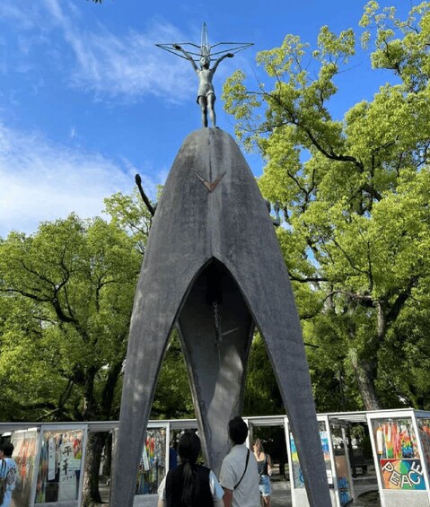 A large metallic sculpture with a figure at the top, set against a backdrop of green trees and a clear sky. 3