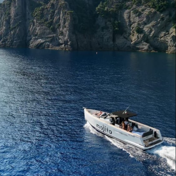 A white boat with 'mojito' written on the side, sailing on a calm sea with rocky cliffs in the background. 1