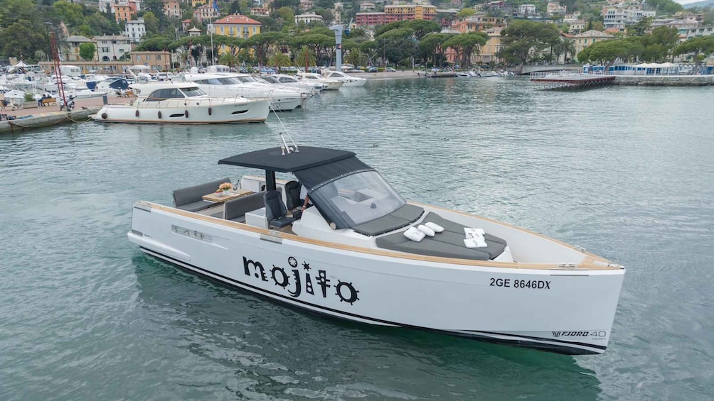 A white boat named 'mojito' with the number '2GF 8646DX' on the side, docked in a marina with other boats and buildings in the background. 3