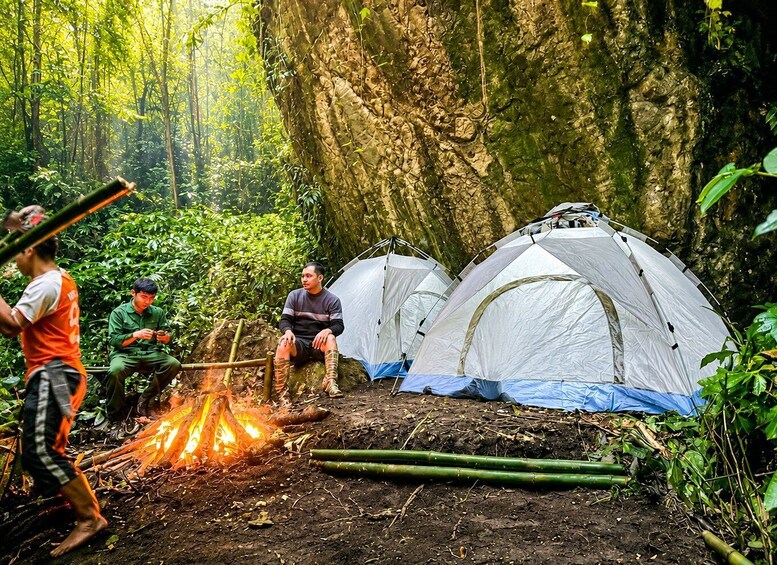 A forest with two tents, a campfire, and people. 1